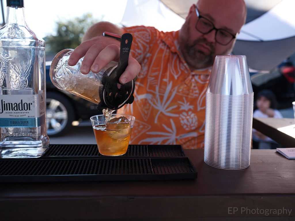 A bartender in an orange Hawaiian shirt pours a cocktail into a glass over ice, with a bottle of tequila and stacks of plastic cups visible on the bar.